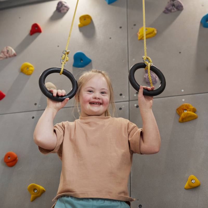 Waist up portrait of cute girl with down syndrome enjoying exercise at home or at sports center and looking at camera while holding at rings against climbing wall, copy space